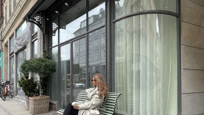 A woman sitting on a bench outside a building with large windows and a green door.