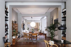 A spacious living area with a dining table, chairs, and a large potted plant. There are shelves with bottles and a staircase leading to another level.