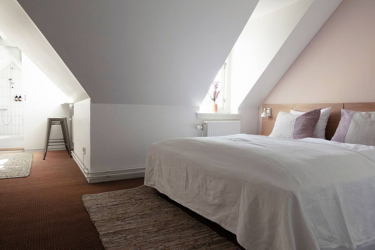 A bedroom with a sloped ceiling, a bed with a white duvet, a radiator, and a window with a view of pink flowers.