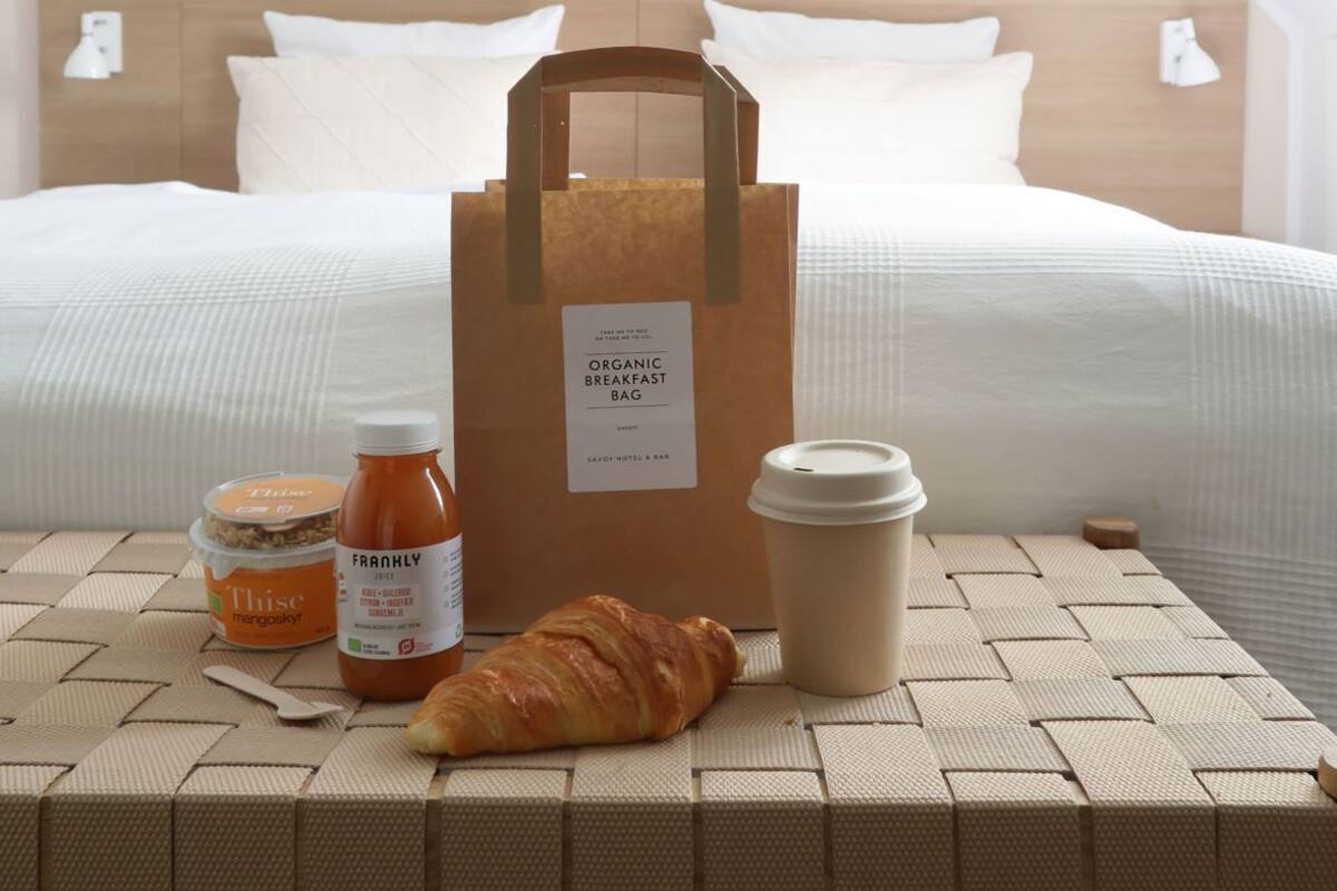 A neatly made bed with a wooden headboard, a breakfast table with a croissant, a bottle of juice, a coffee cup, and an organic breakfast bag.