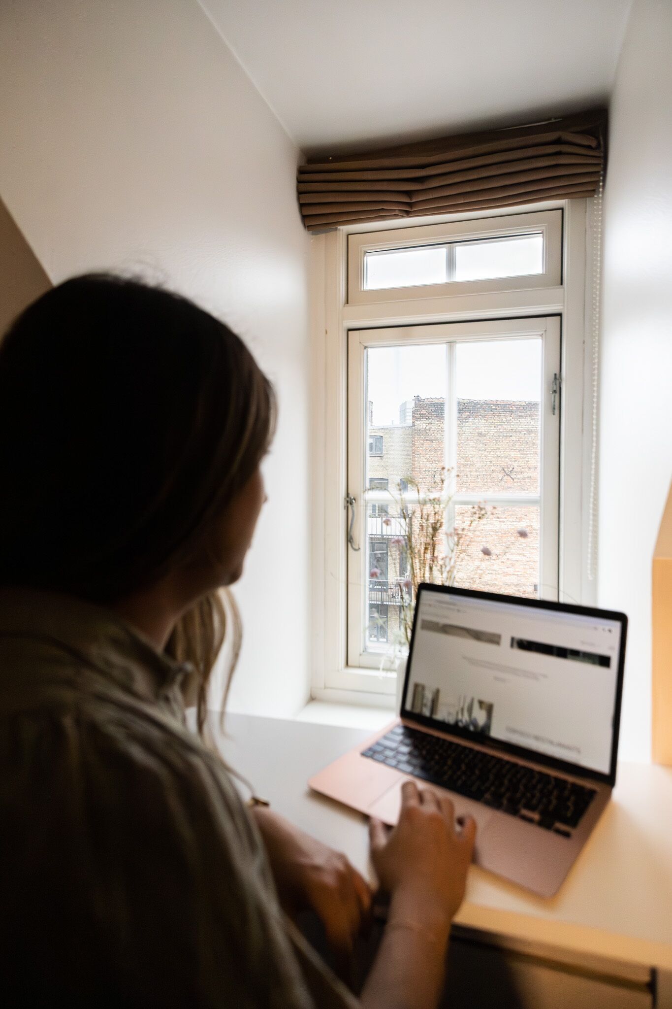 Una persona usando una computadora portátil en un escritorio junto a una ventana con vistas a edificios.