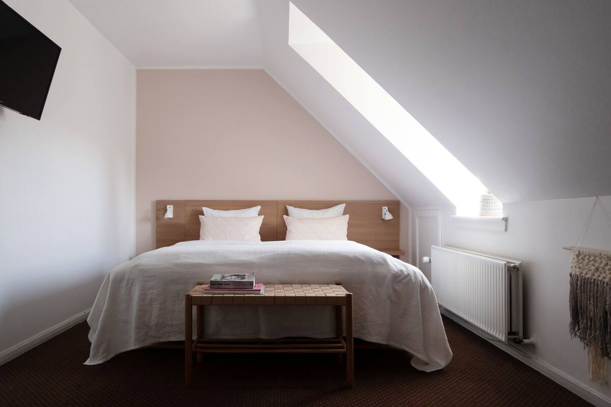 A bedroom with a sloped ceiling, a bed with white bedding, a bedside table with a book, and a radiator.