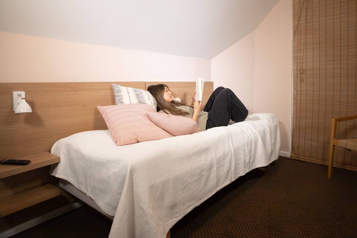 A single bed with a wooden headboard, a bedside table with a lamp, and a chair in a hotel room.