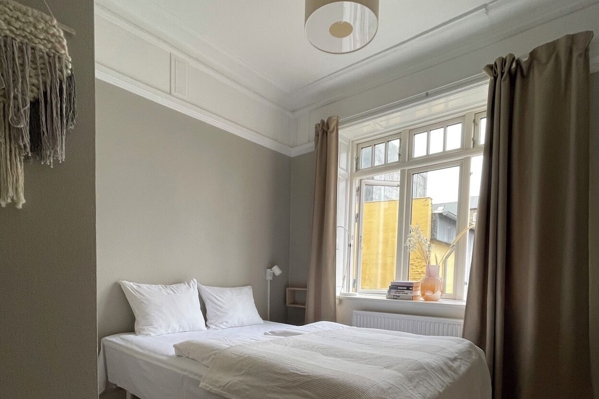 A bedroom with a bed, a window with curtains, a hanging light fixture, and a radiator.