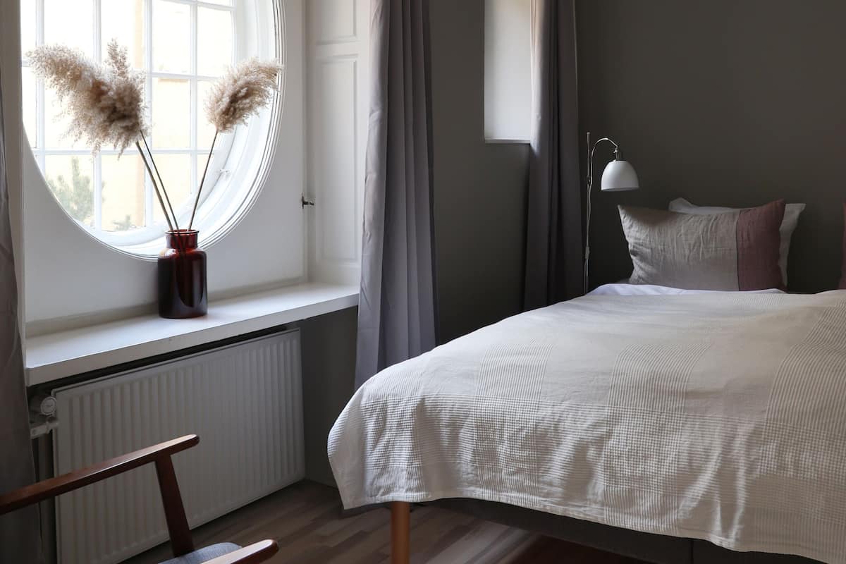 A bedroom with a large bed, a window with curtains, a bedside lamp, and a chair.