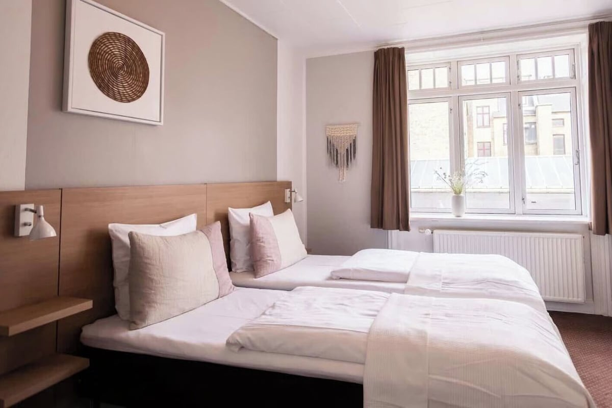 A hotel room with two beds, a bedside table, a lamp, a window with curtains, and a wall-mounted decor piece.