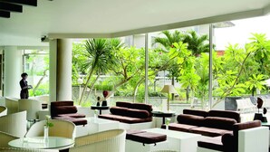 A modern indoor lounge with wicker furniture, large windows, and tropical plants.