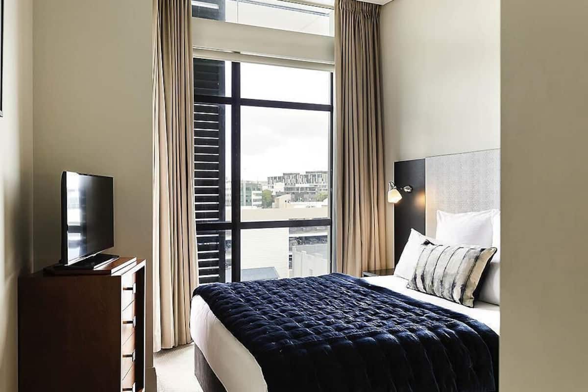 A hotel room with a bed, a TV on a stand, a window with curtains, and a city view.