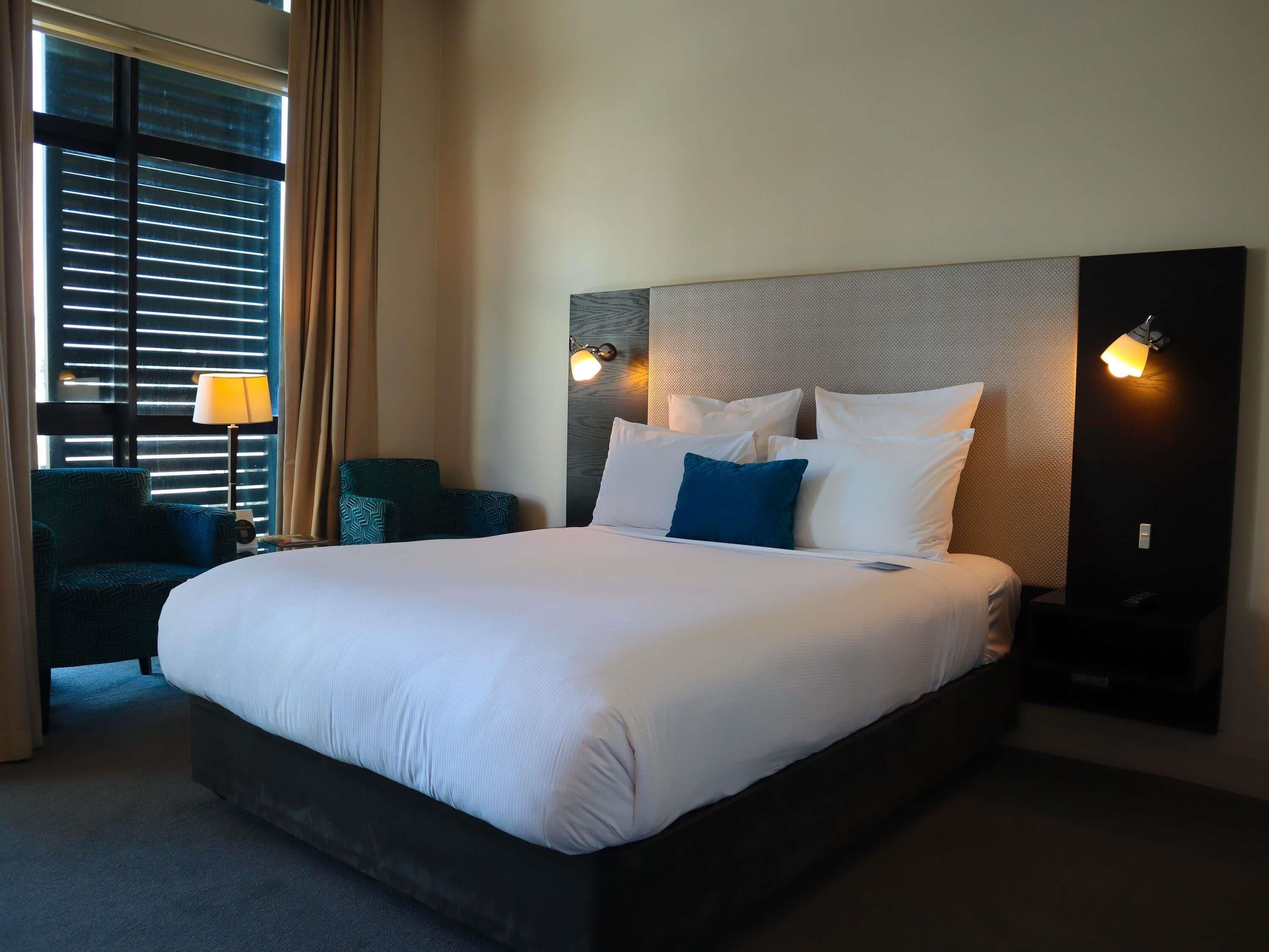 A hotel room with a large bed, two bedside tables, a chair, and a window with blinds.