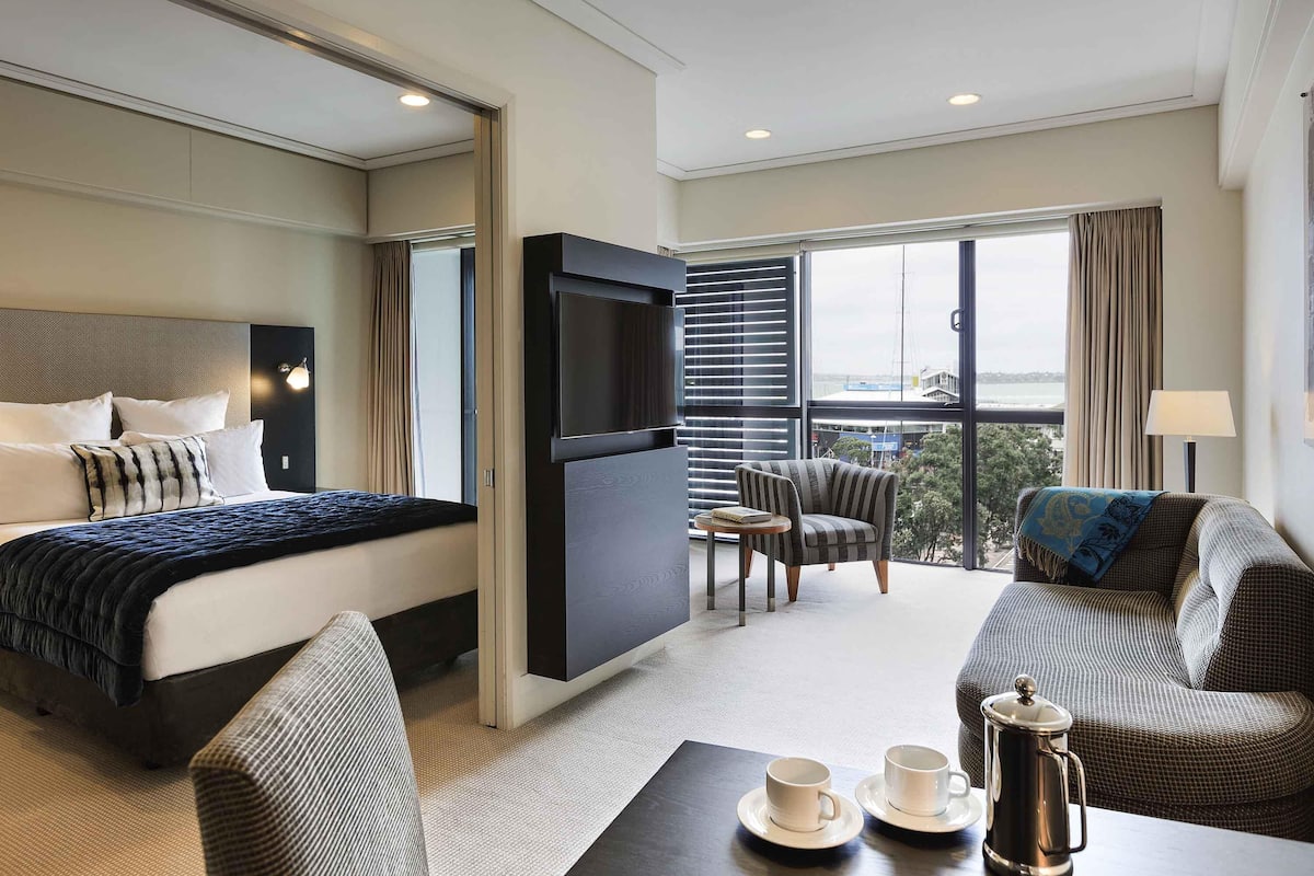A hotel room with a bed, a TV, a sofa, a table with cups and a coffee pot, and a view of the city through large windows.