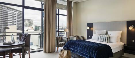 Habitación de hotel con una cama grande, un escritorio con silla, una ventana con cortinas y vistas a la ciudad.