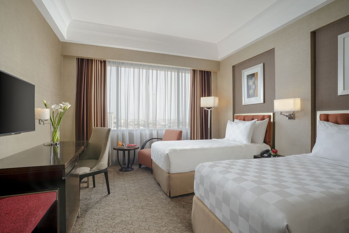 Ein Hotelzimmer mit Schreibtisch, zwei Betten, Fernseher, Sessel, kleinem Tisch und einem Fenster mit Vorhängen.