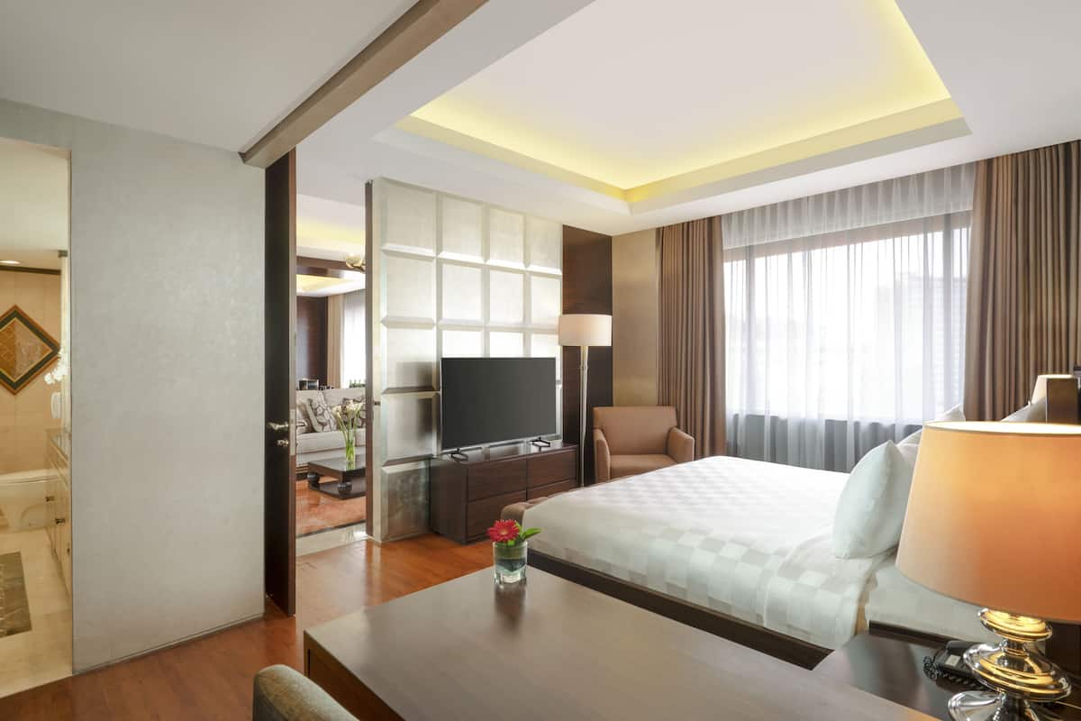 Ein Hotelzimmer mit einem Bett, einem Fernseher, einem Schreibtisch mit Lampe und Blick auf die Stadt durch große Fenster.