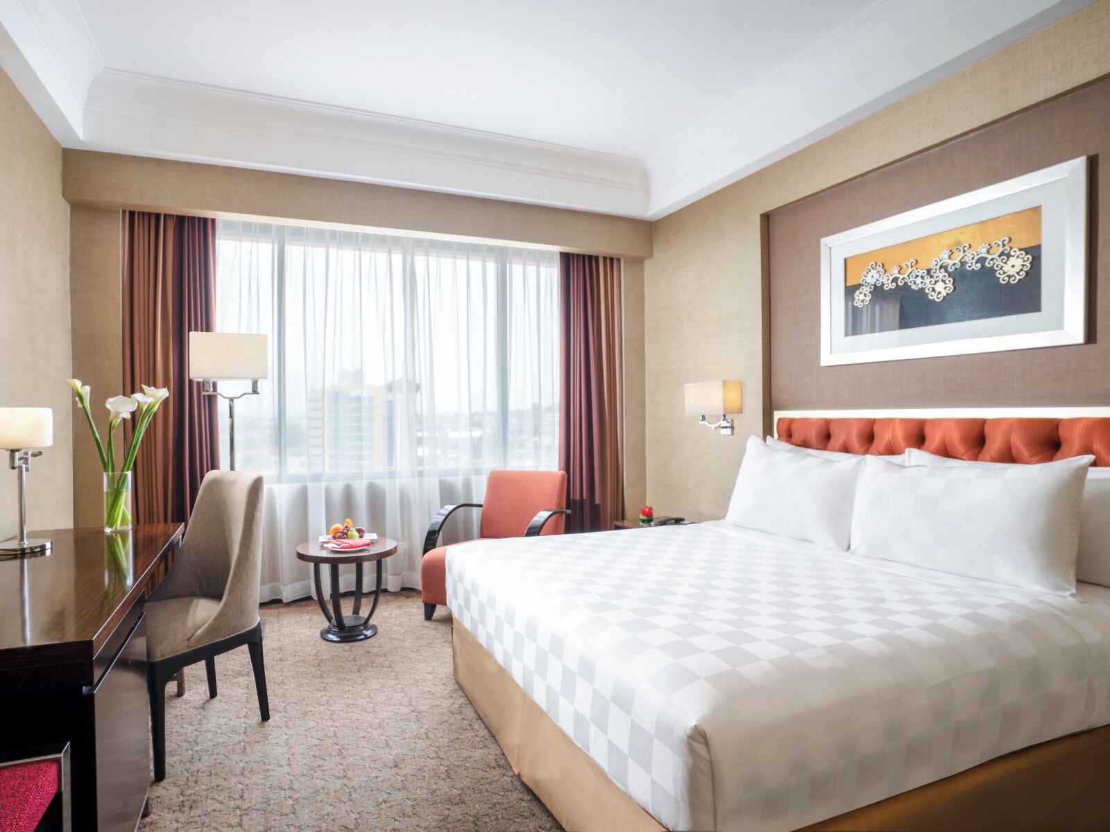 A hotel room with a large bed, two chairs, a desk with a lamp, a small table with fruit, and a window with curtains.