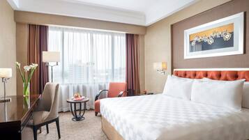 A hotel room with a large bed, two chairs, a desk with a lamp, a small table with fruit, and a window with curtains.
