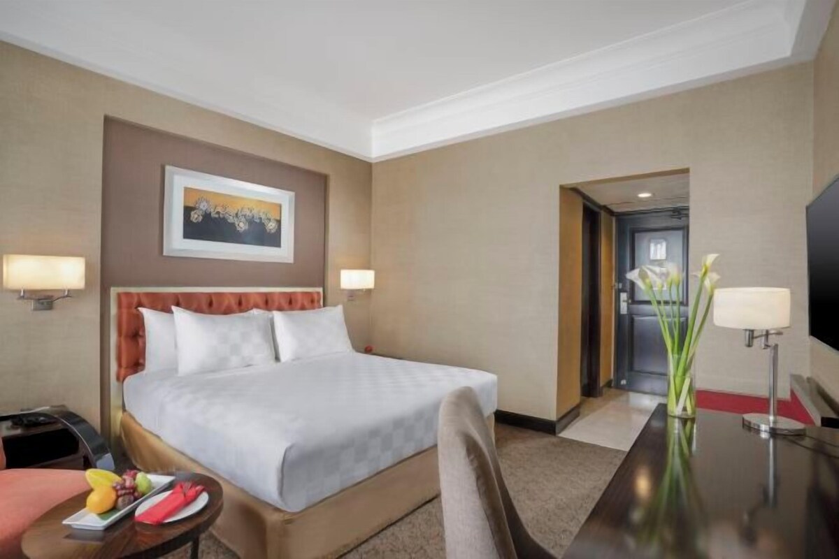 A hotel room with a bed, bedside lamps, a desk, a chair, a small table with fruit, and a vase with flowers.
