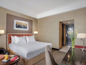 Ein Hotelzimmer mit einem Bett, Nachttischlampen, einem Esstisch mit Obst, einer Vase mit Blumen und einem Bild an der Wand.