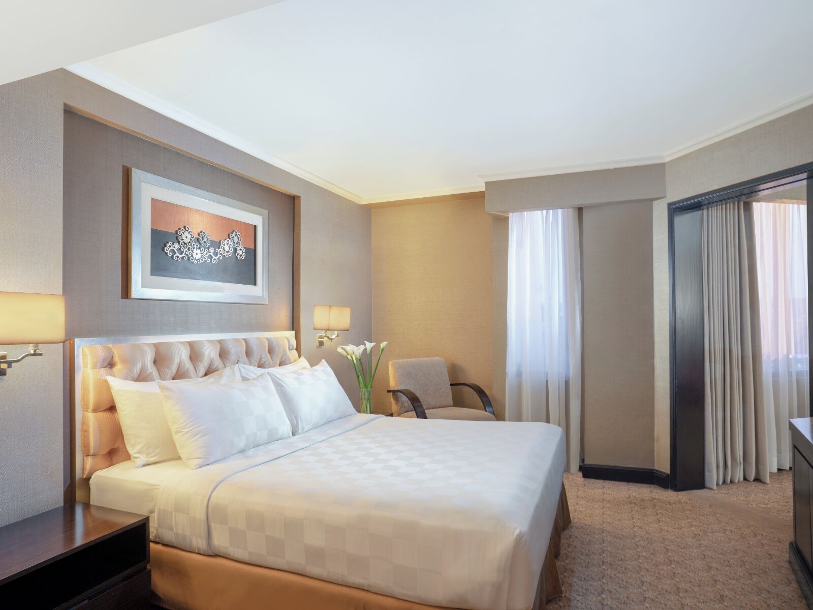 Una habitación de hotel con una cama grande, un escritorio, una silla, una lámpara y un ventanal con cortinas.