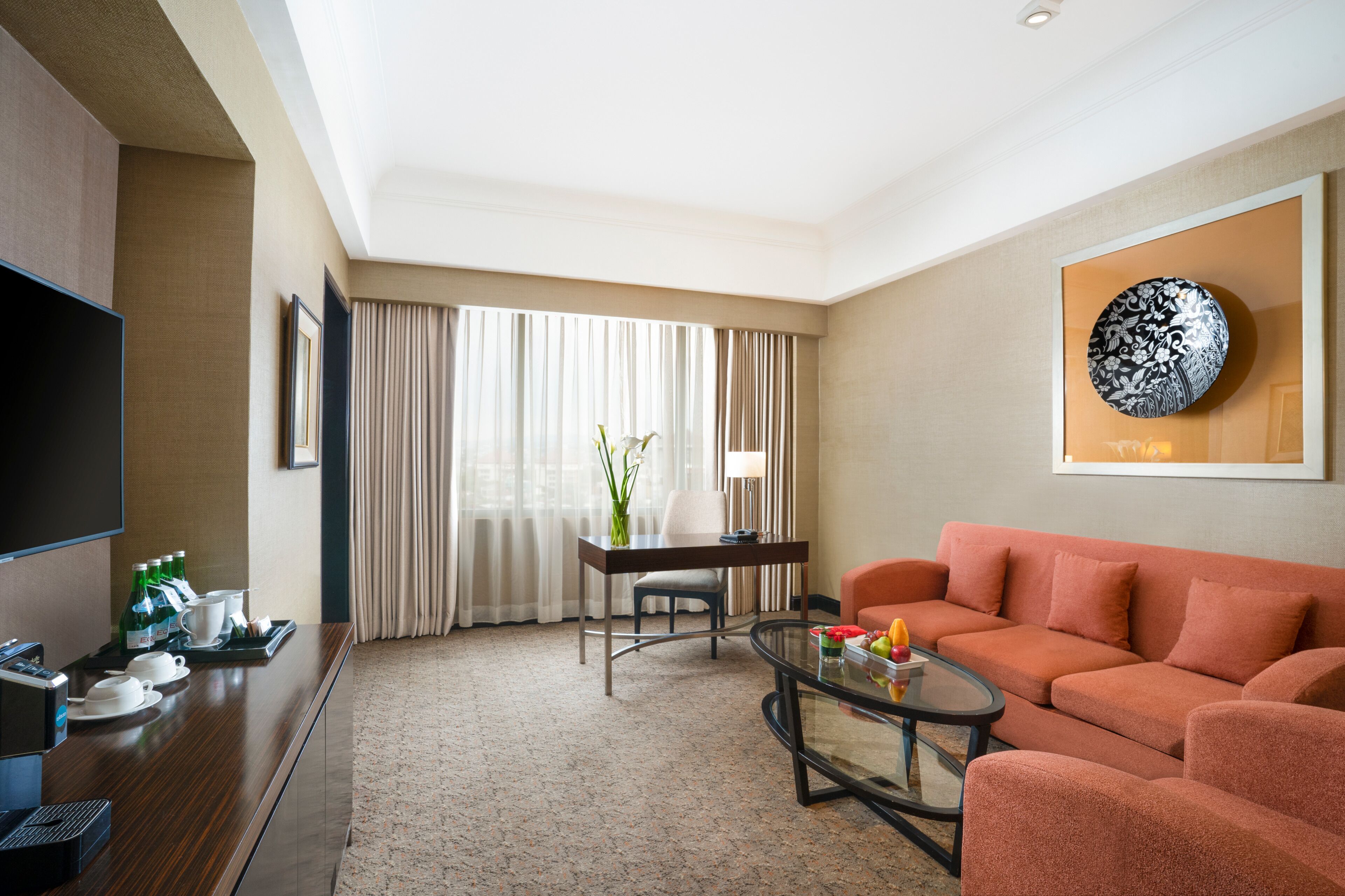 Ein Hotelzimmer mit Fernseher, einer Couch, einem Couchtisch, einem Esstisch mit Stühlen und einem großen Fenster mit Vorhängen.