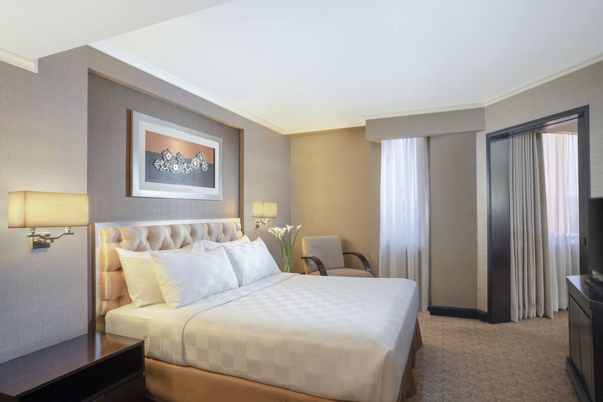 Ein Hotelzimmer mit einem großen Bett, zwei Nachttischen, einem Sessel, einem Fernseher und einem Bild an der Wand.