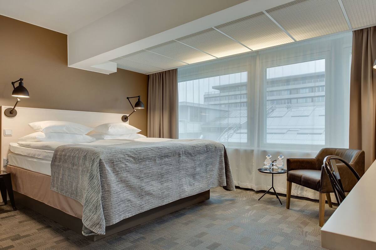 Ein Hotelzimmer mit Bett, Nachttischlampen, einem Sessel, einem kleinen Tisch mit Flaschen und Blick auf Gebäude durch große Fenster.