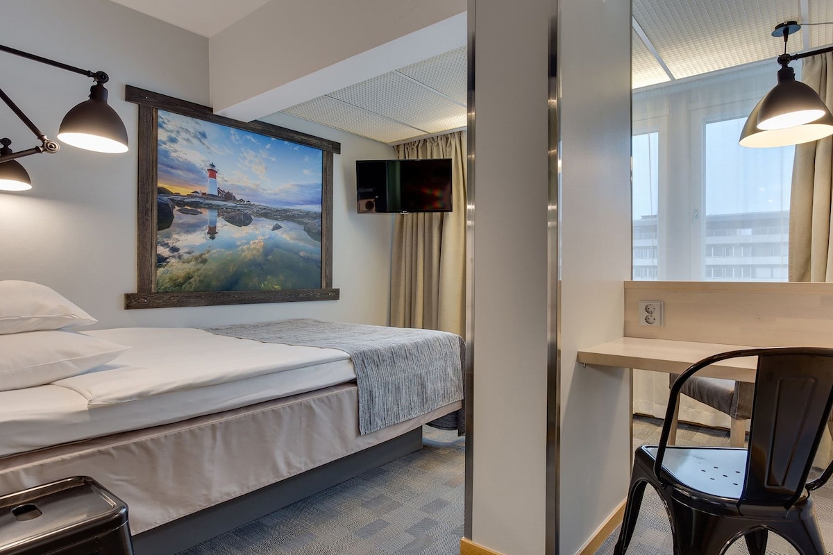 Habitación de hotel con cama, escritorio, silla, televisor y un cuadro de un faro.