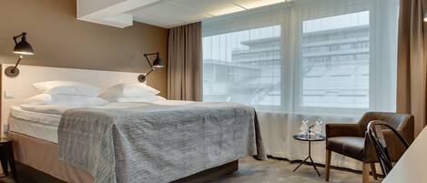 A hotel room with a bed, a chair, a small table, and a window with a view of buildings.