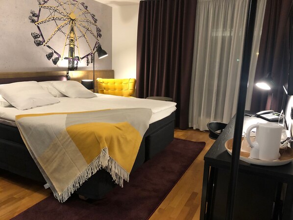 A hotel room with a bed, a desk with a lamp, a coffee cup, and a wall decoration.