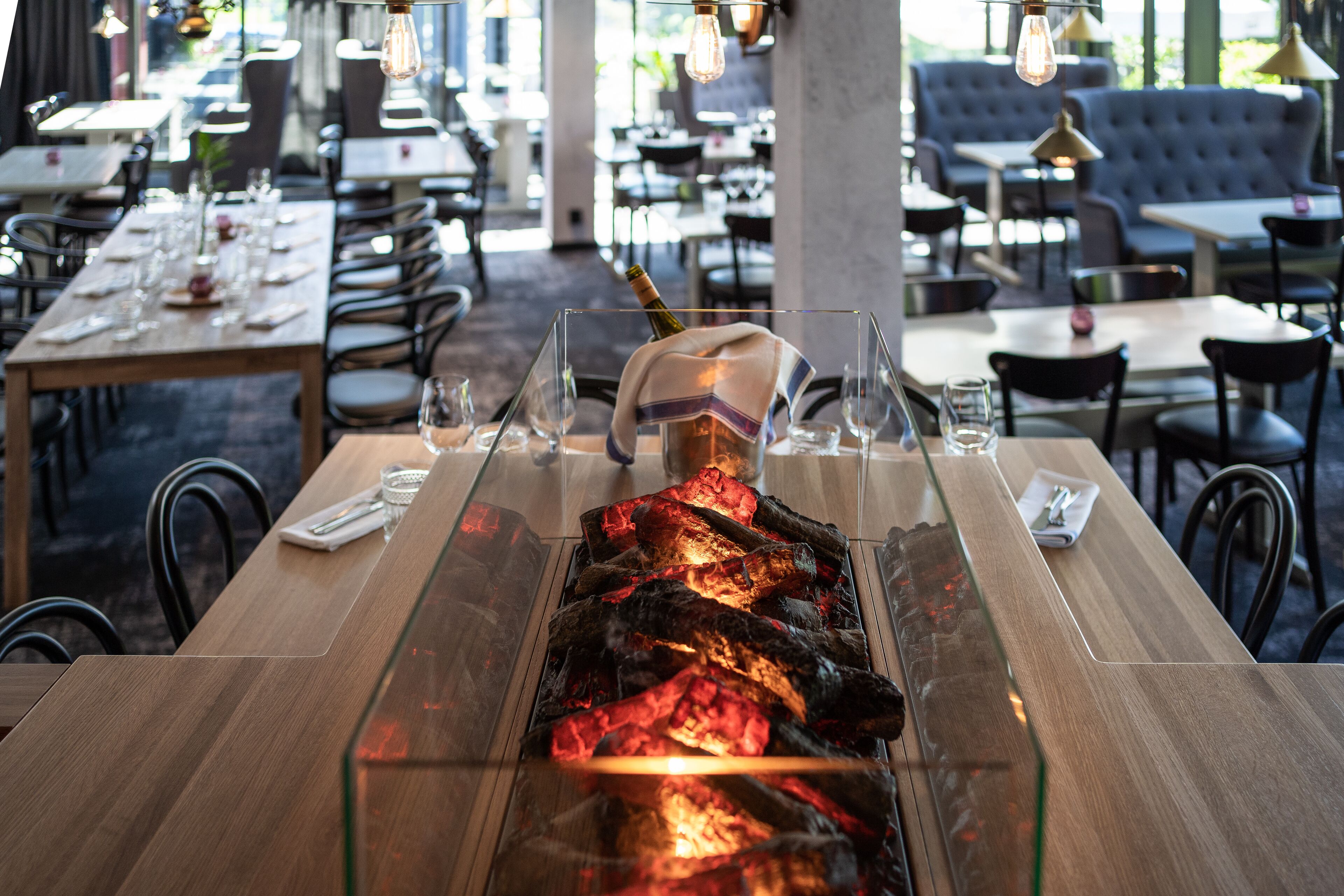 A restaurant with a long wooden table, a glass-top table with a lit charcoal fire, and a cozy seating area in the background.