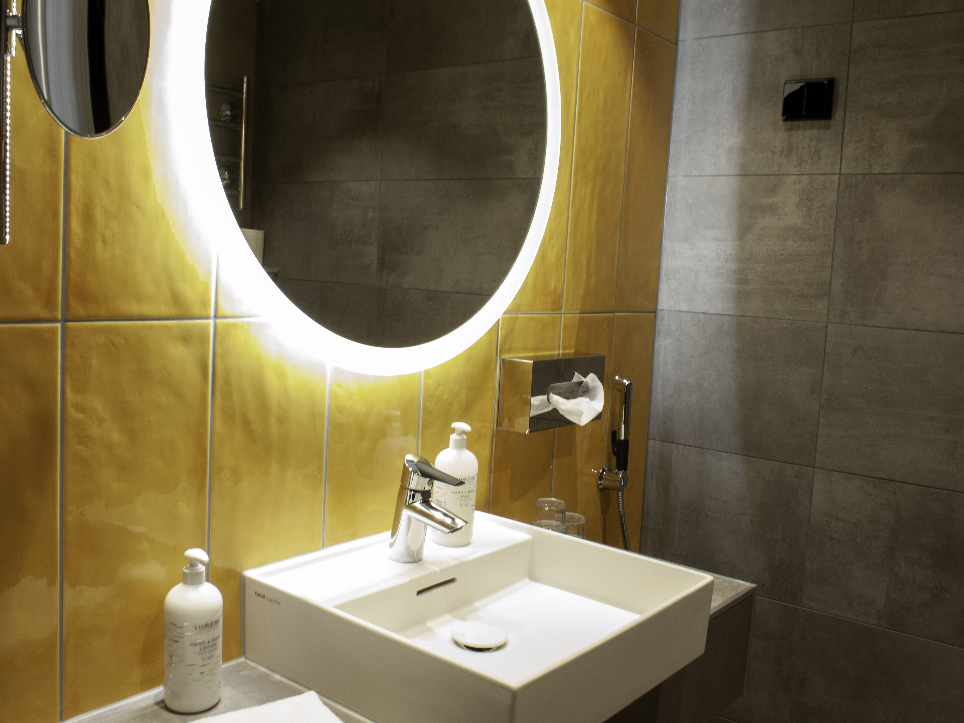 A modern bathroom with a white sink, a mirror with a circular light, and a towel rack.