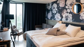 Quarto de hotel com cama, escrivaninha, cadeira, janela grande e papel de parede floral.