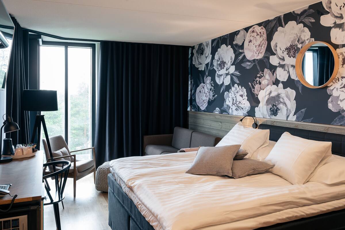 A hotel room with a bed, a desk, a chair, a large window, and a floral wallpaper.