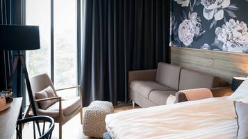 Habitación de hotel con cama, sofá, sillón, pequeño reposapiés, escritorio con lámpara y un ventanal con cortinas.