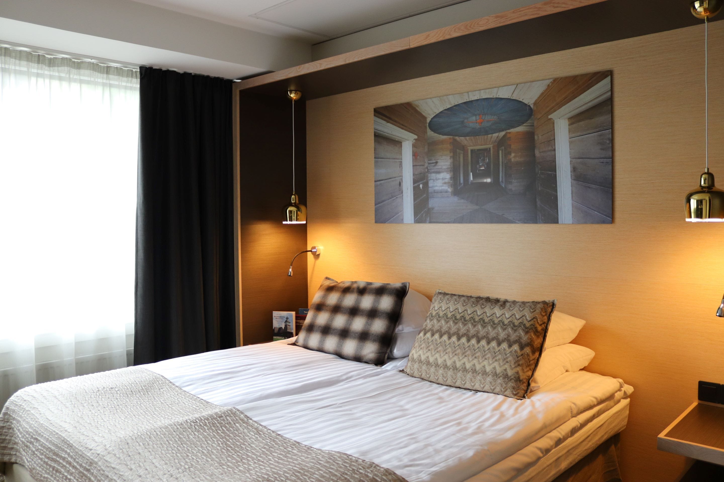 A neatly made bed with a checkered pillow and a zigzag pillow, a wall-mounted painting of a hallway, and two pendant lights.