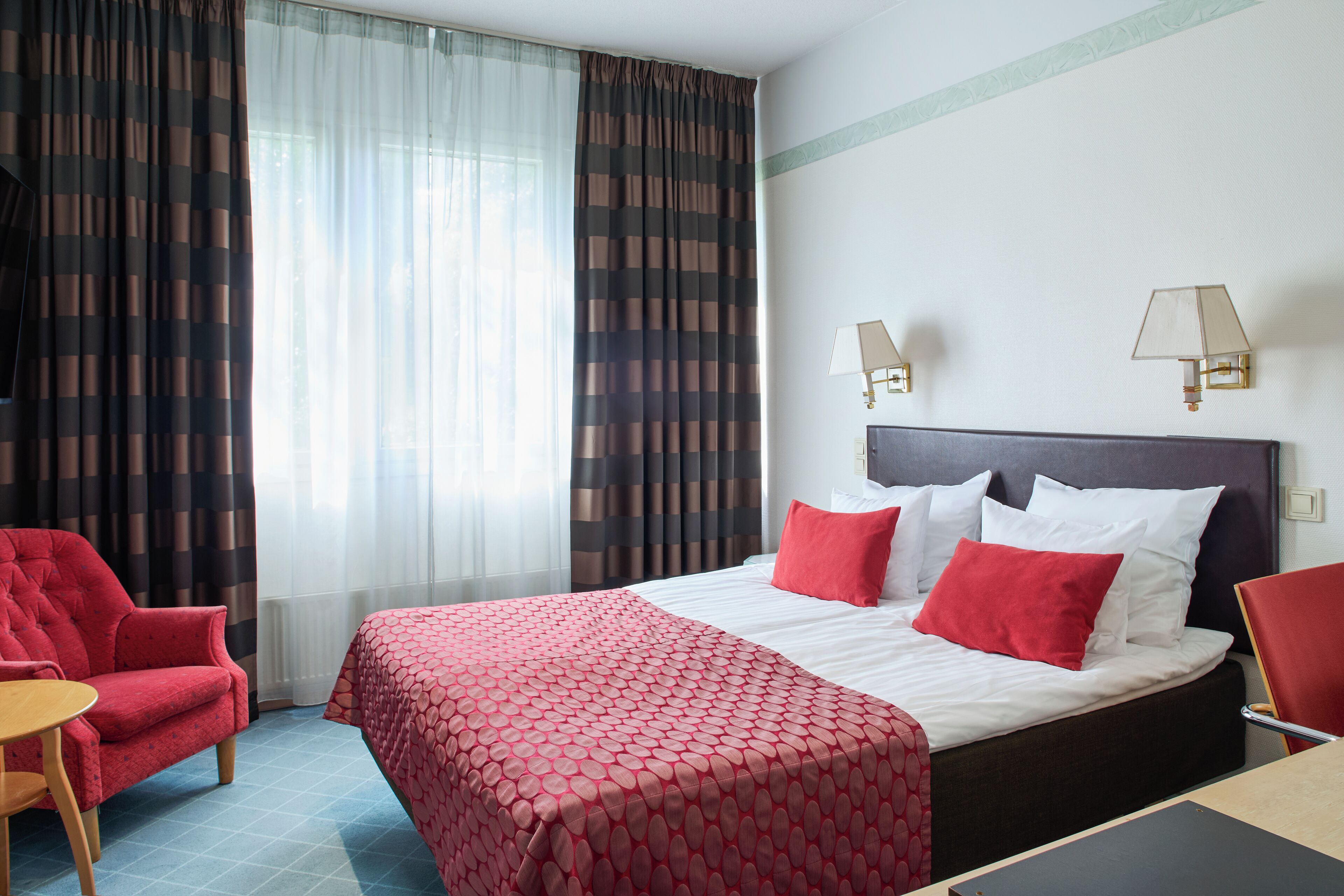 Una camera d'albergo con un letto, due abat-jour, una poltrona rossa, un tavolino e una finestra con tende.