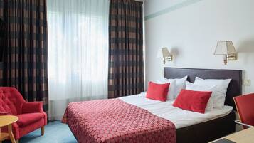 Una camera d'albergo con un letto, due abat-jour, una poltrona rossa, un tavolino e una finestra con tende.