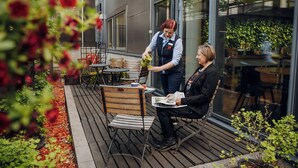 An outdoor seating area with wooden chairs and tables, a person pouring a drink, and another person reading a magazine. - Original Sokos Hotel Alexandra (Jyväskylä)