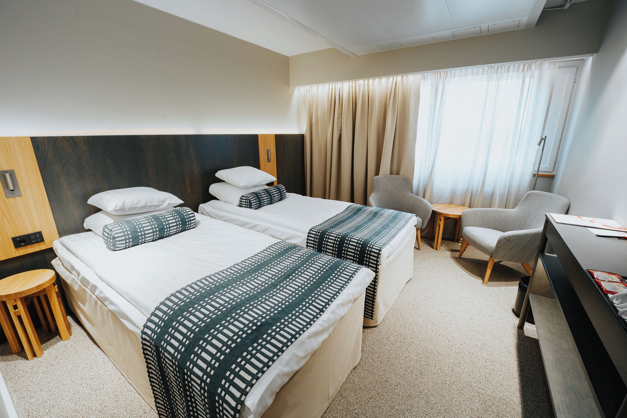 A hotel room with two beds, a desk, a chair, and a small stool.