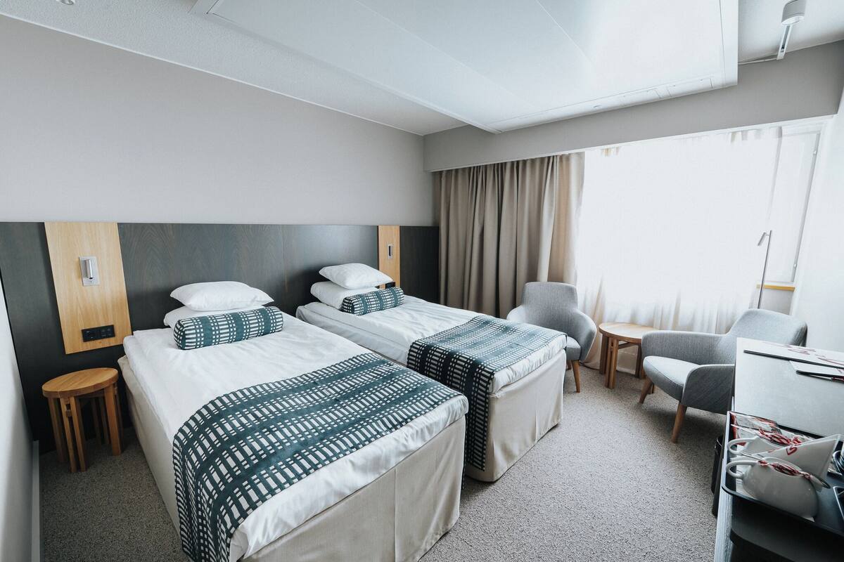 A hotel room with two beds, a desk, a chair, and a small stool.