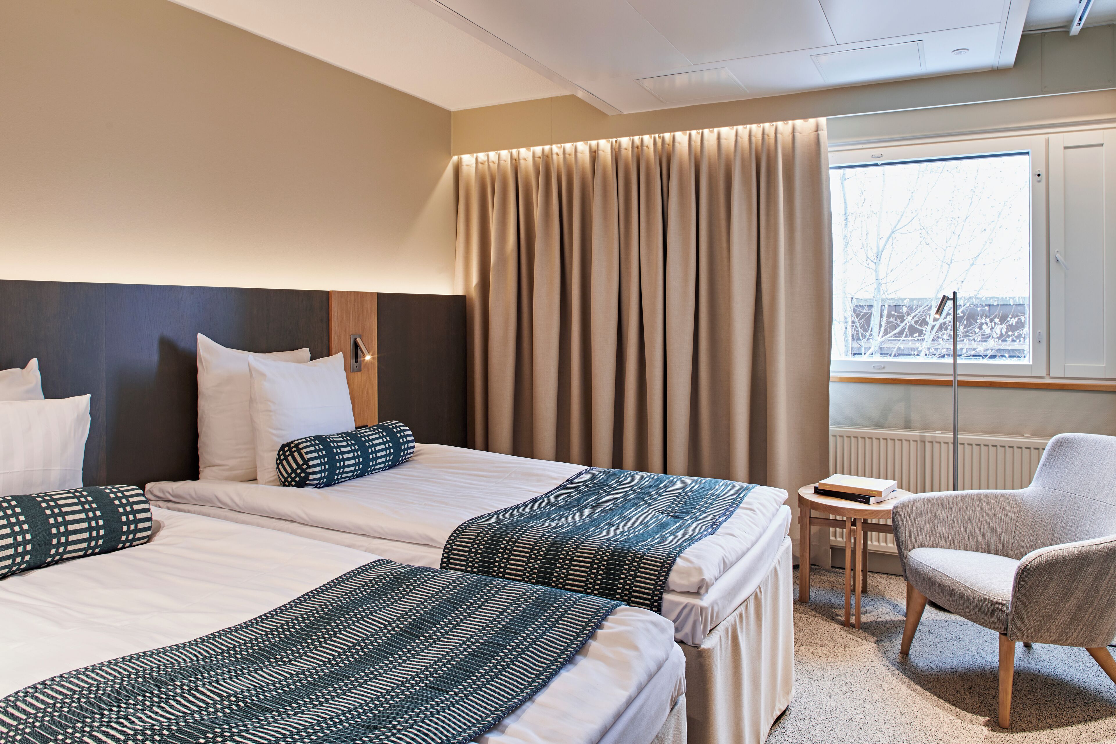 Une chambre d’hôtel avec deux lits, une chaise, une petite table et une fenêtre avec des rideaux.