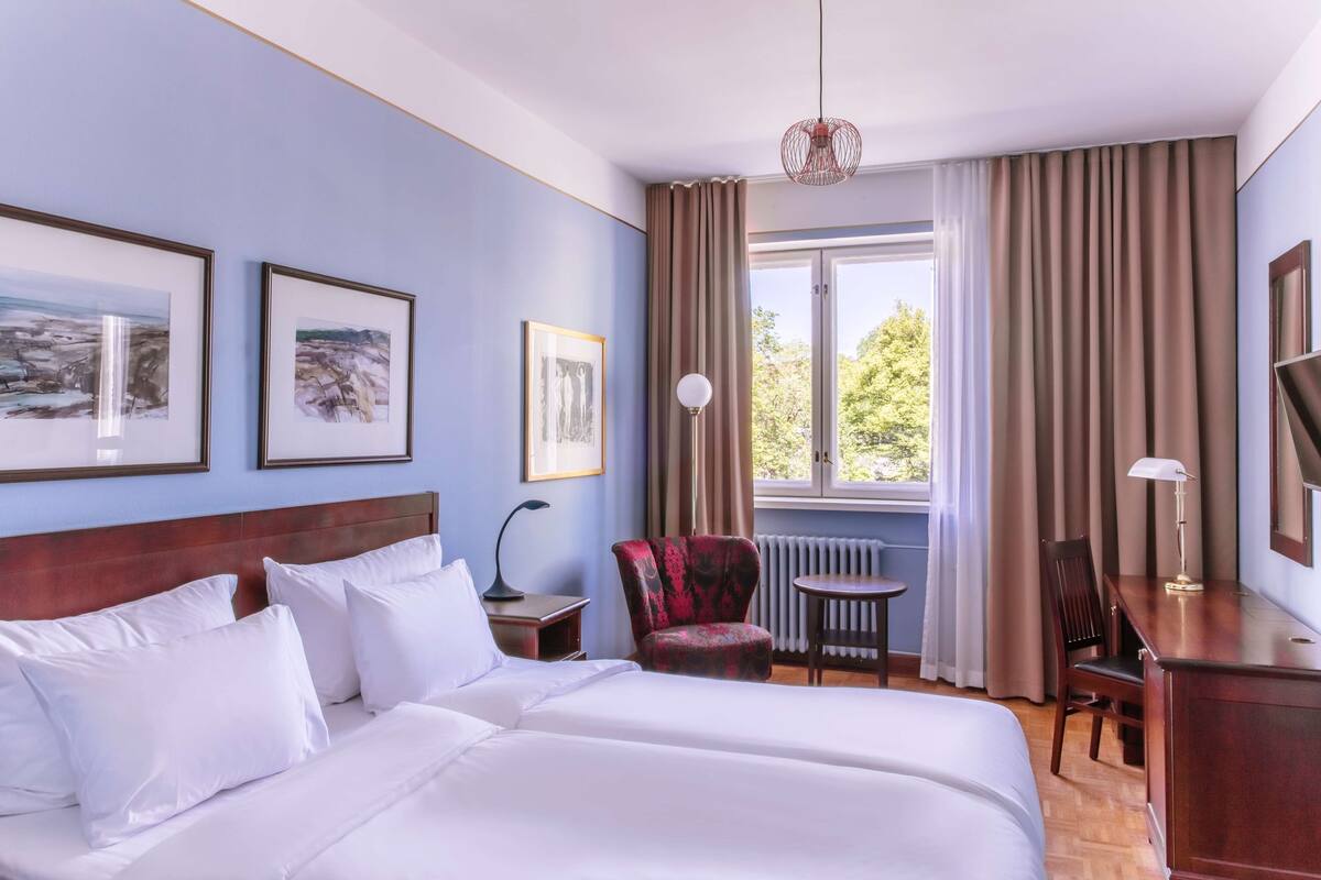 Habitación de hotel con una cama grande, dos cuadros en la pared, un sillón rojo, una mesita y un escritorio con una lámpara.