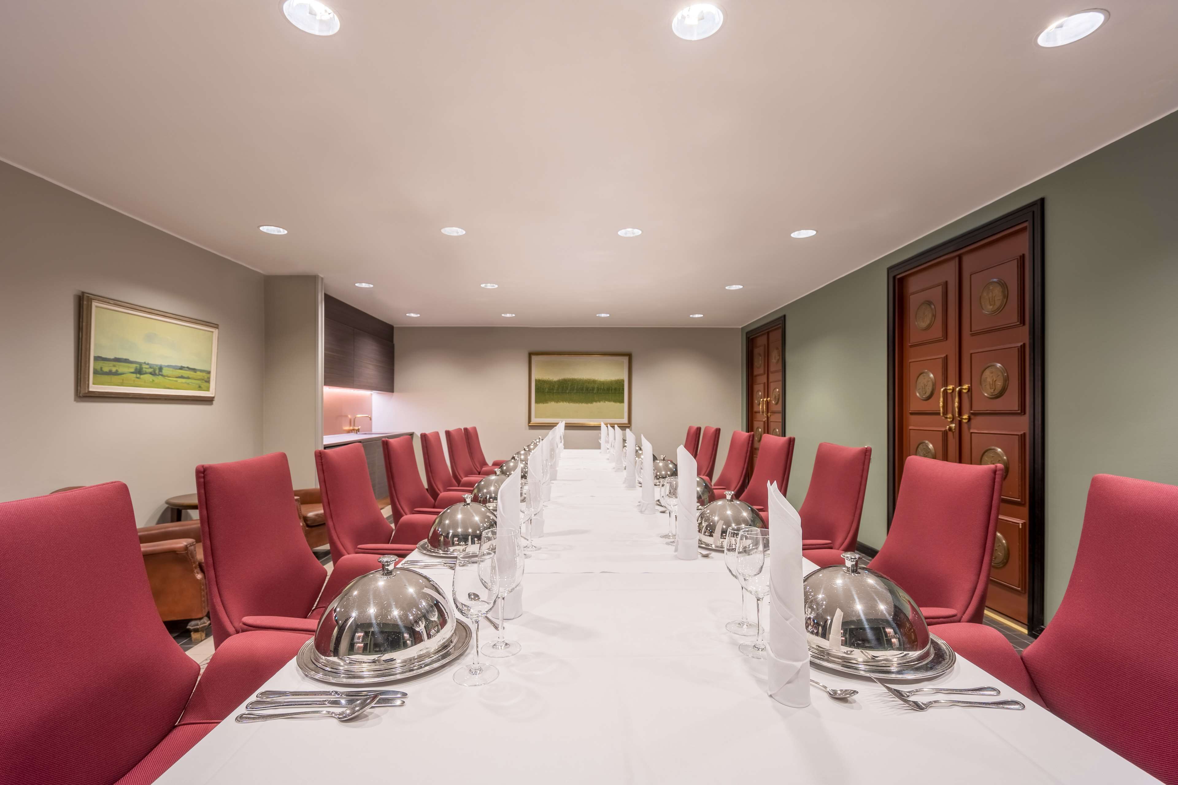 Un salón de conferencias con una larga mesa dispuesta para un evento formal, rodeado de sillas rojas, y una pintura en la pared.