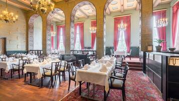 A formal dining room with multiple tables set for service, ornate chandeliers, and a red carpeted floor.