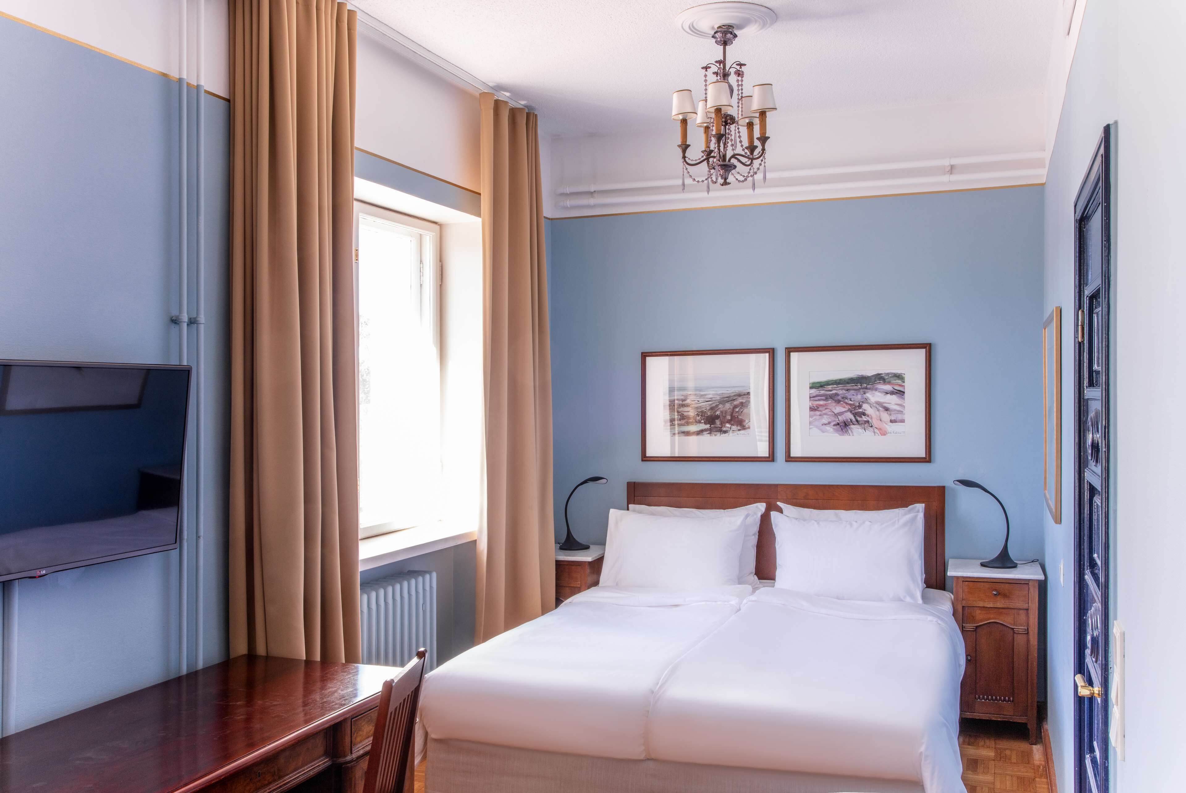 A hotel room with a bed, a desk, a chair, a TV, and two framed pictures on the wall.