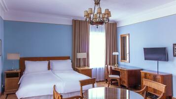 A hotel room with a double bed, a desk with a chair, a television, and a chandelier.