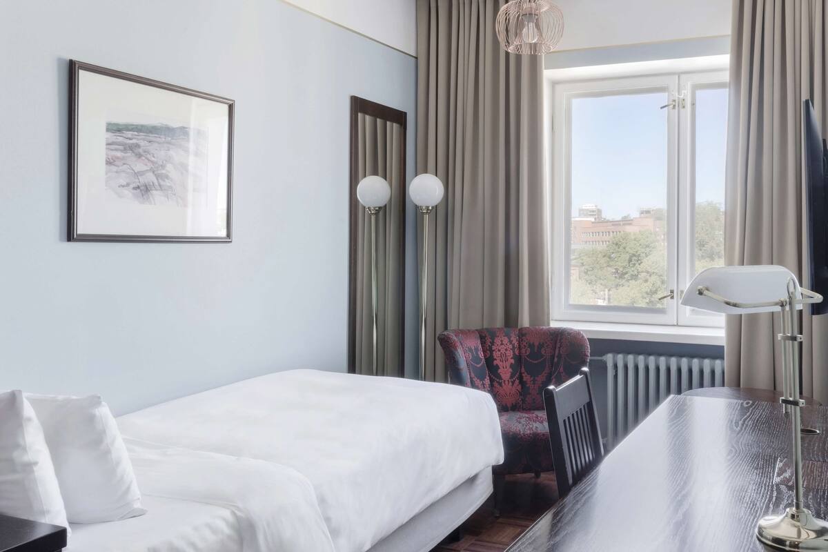 Habitación de hotel con cama, una silla, un escritorio, una lámpara, un cuadro en la pared y una ventana con cortinas.