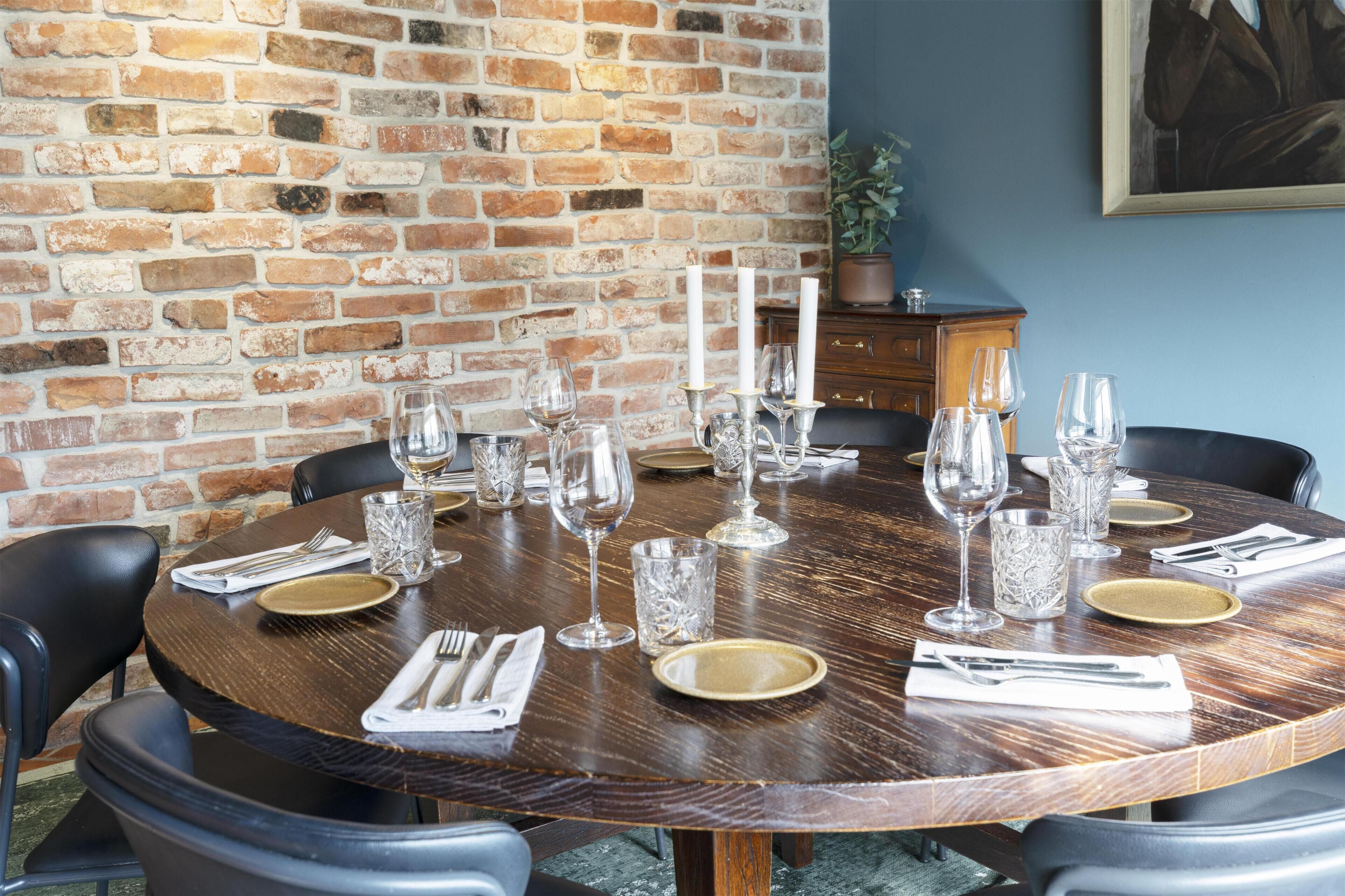 Une table de salle à manger ronde, mise avec des assiettes, des couverts et des verres à vin, placée devant un mur de briques et sur un fond bleu.