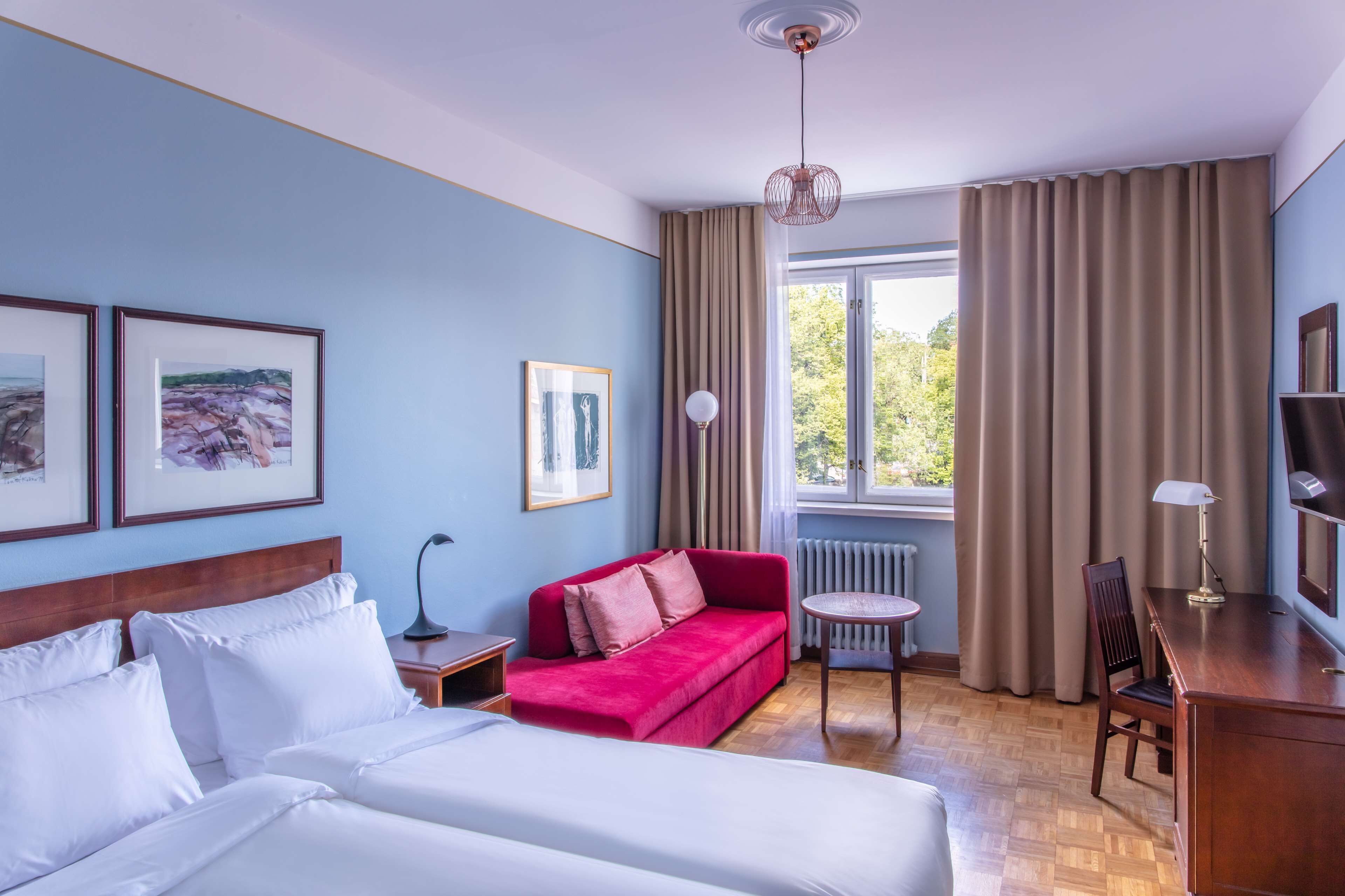 Quarto de hotel com cama, sofá vermelho, escrivaninha com luminária e quadros nas paredes.