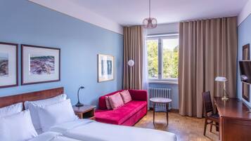 Quarto de hotel com cama, sofá vermelho, escrivaninha com luminária e quadros nas paredes.