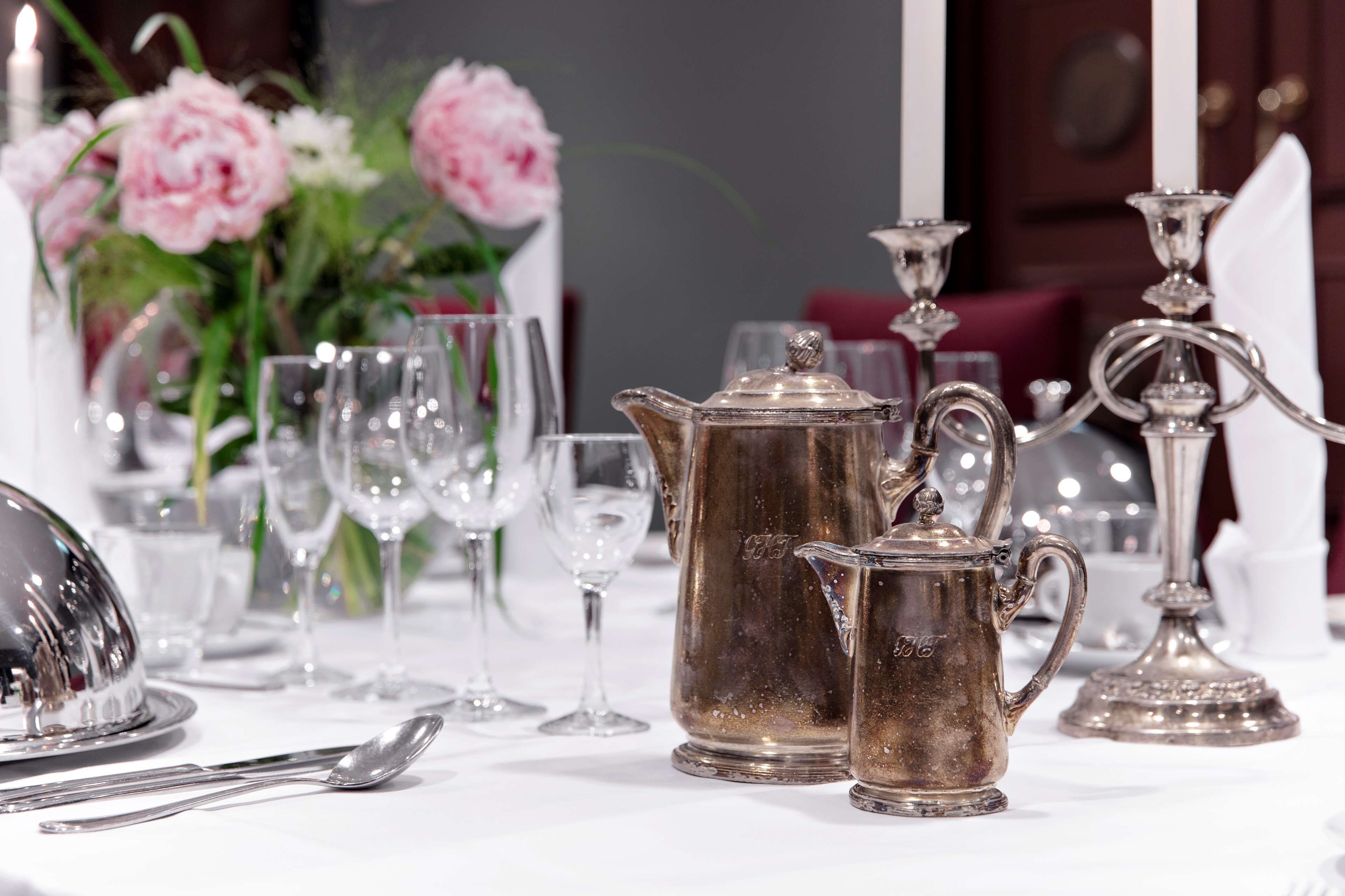 Una mesa dispuesta con cubiertos, una tetera plateada y una jarra plateada, con flores rosadas al fondo.